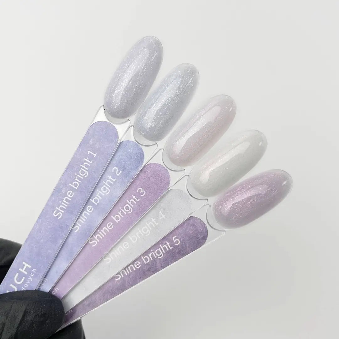 A hand wearing a black glove holds five nail polish color swatch sticks labeled Shine bright 1 through Shine bright 5 on a plain light background. The swatches show five different pastel shimmer shades: silvery white, pale lavender, soft pink, milky white, and light lilac.
